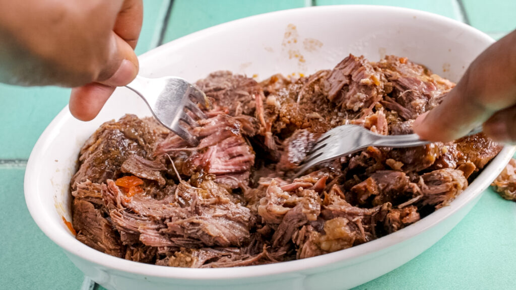 shredding the birria meat with 2 forks. the cooked meat is in a white oval dish