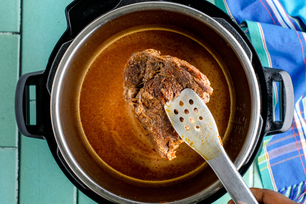 overhead image of a piece of cooked chuck roast being lifted out of an instant pot. there is a red sauce in the background
