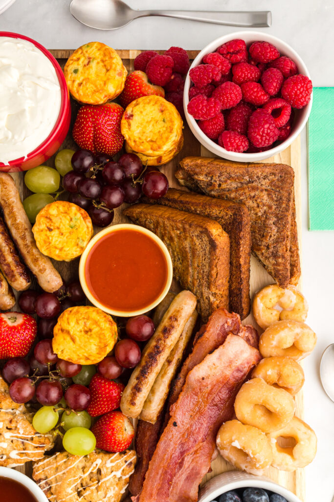 overhead shot of breakfast items on a wooden board with berries, syrup, sausage, bacon, french toast, fruit dip and mini donuts