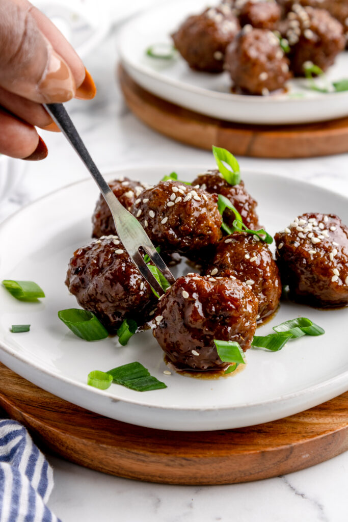 plate of meatballs topped with scallion and sesame seeds. One meatball is being picked up with an appetizer fork