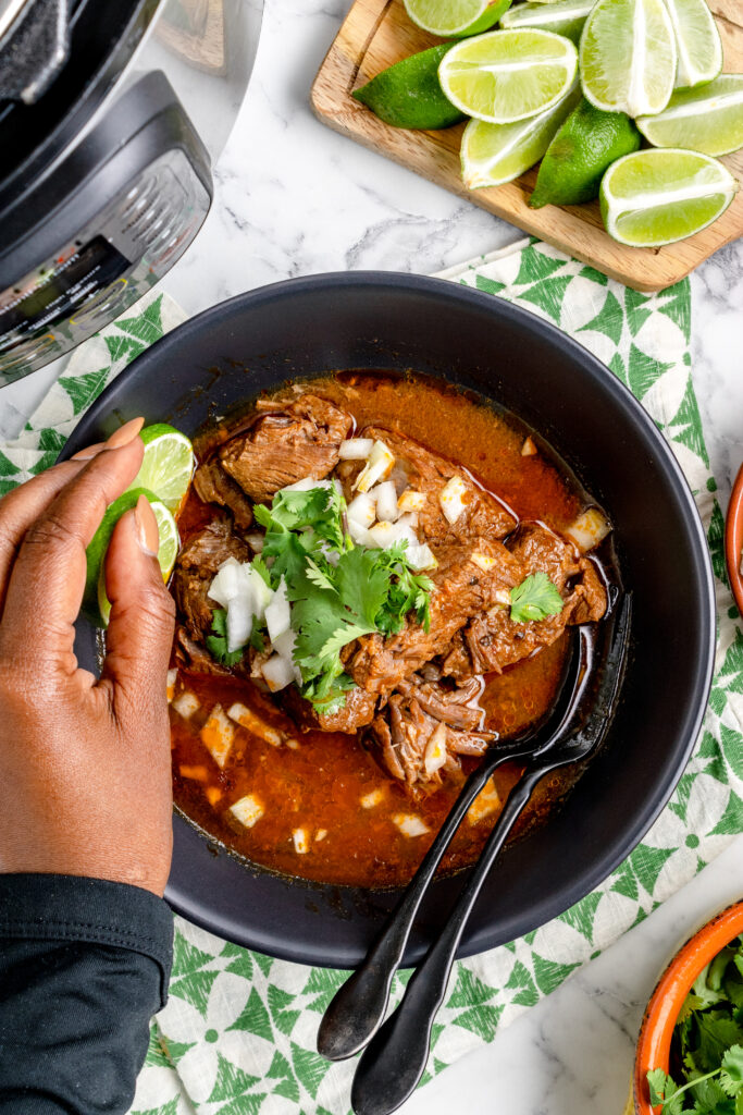birria stew in a bowl overhead shot with female hand garnishing with lime slices