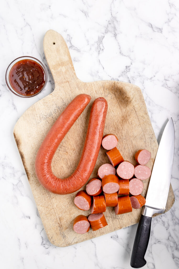 whole kielbasa and sliced kielbasa on a wooden cutting board with a knife and a small bowl of bbq sauce